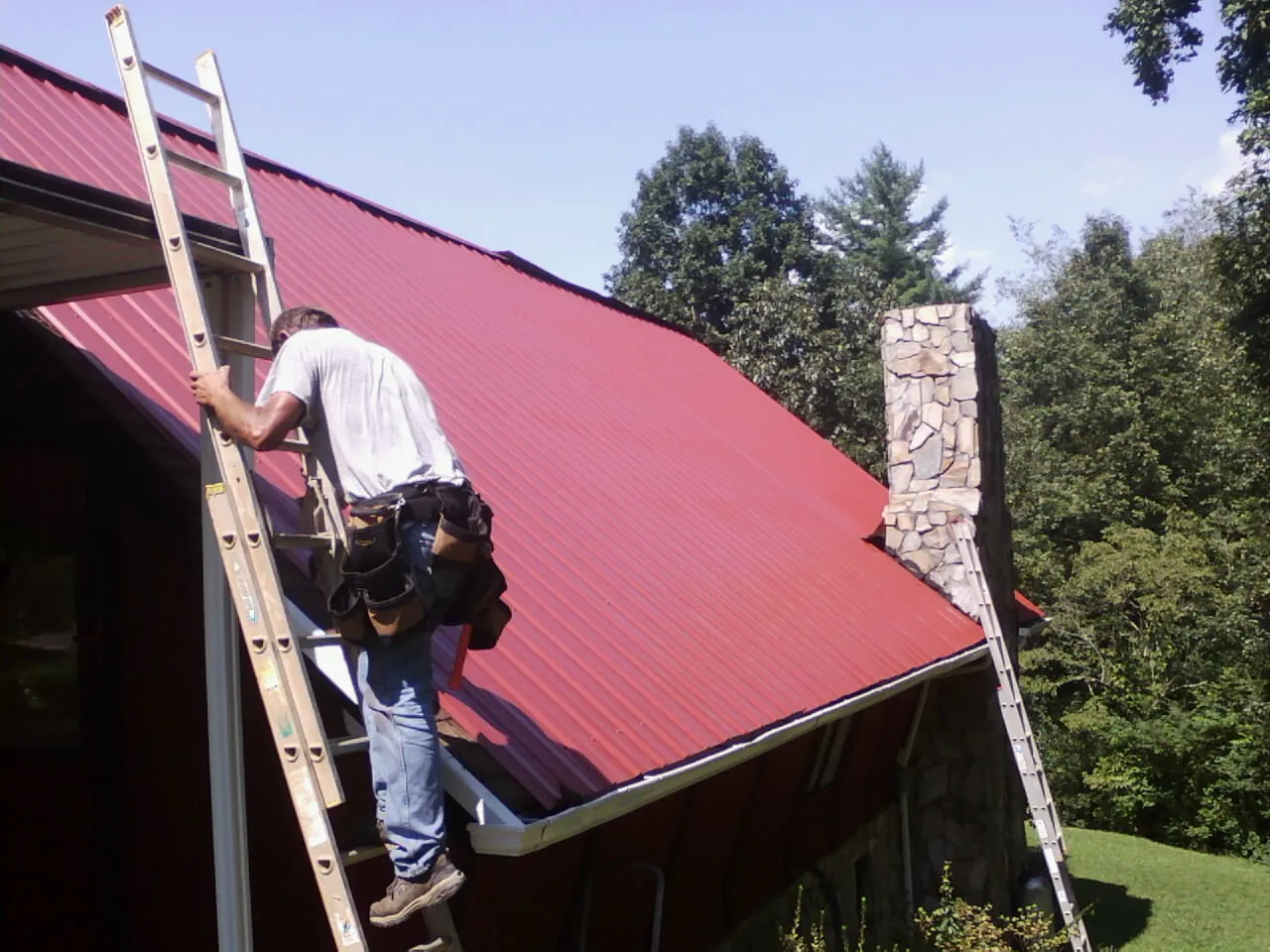 Roofer on a ladder beside a red metal roof and stone chimney — Soffit & Fascia Repair by Roofing Brothers in Changewater